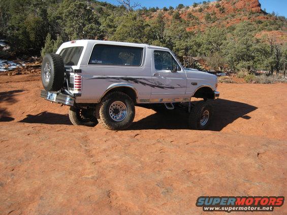 White 1996 Ford Bronco with aftermarket wheels, spare tire on rear, parked on red dirt