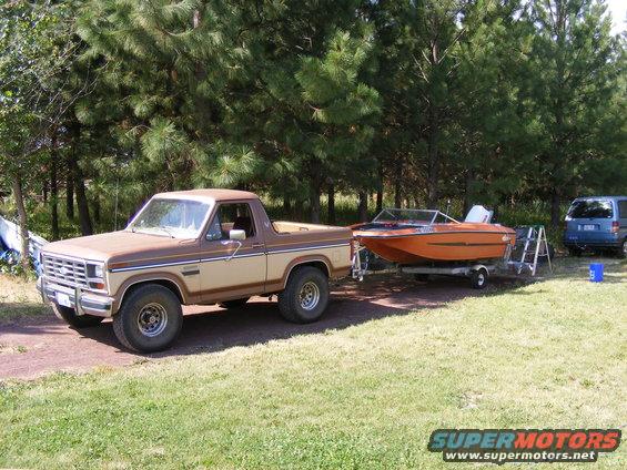Brown and tan Ford Bronco towing an orange boat on a trailer. Blue SUV parked to the right
