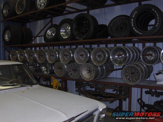 shelving unit filled with tires and car wheels, part of a white car hood in foreground