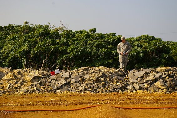 Soldier in camouflage uniform stands on a pile of rubble and asphalt with green foliage behind him