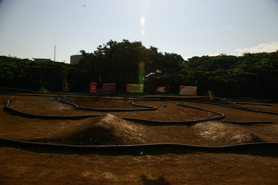 RC car track with jumps and banked turns. A car is airborne over a jump. People stand near sponsor banners