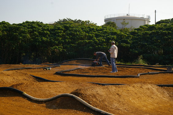 Remote control cars race on a dirt track with two people nearby and a water tower in the background