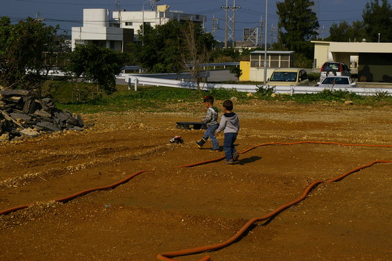 Two boys walk on a dirt track with a remote control car; orange hose visible