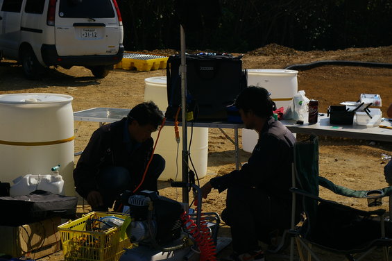 Two men crouch near equipment and barrels outdoors, with a white van in the background