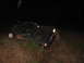 1984 Ford Bronco II - A muddy black Ford Bronco II is partially submerged in dirt and vegetation, its headlights illuminated in the darkness.