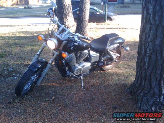 Black motorcycle parked on grass next to two trees. A car is visible in the background