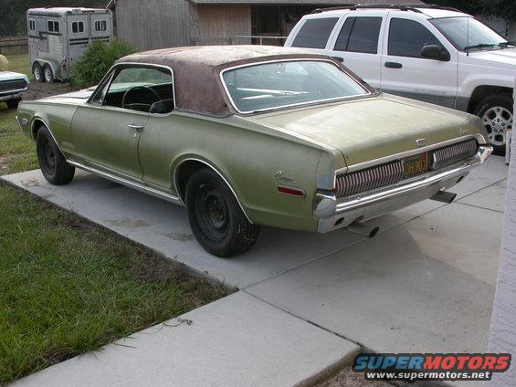 Olive green 1967 Mercury Cougar with a brown vinyl top, parked on a concrete driveway next to a white SUV