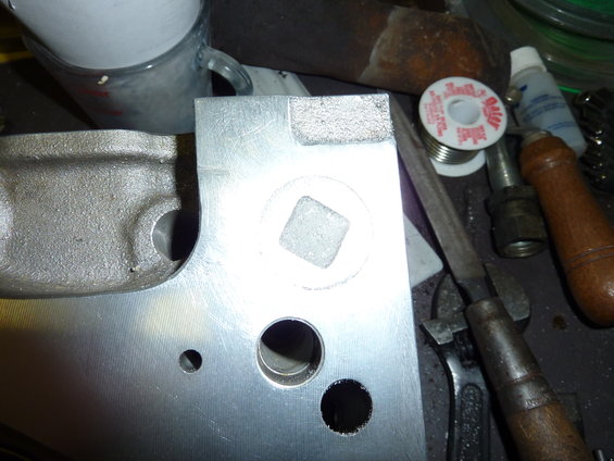 Metal part with a square recess and circular holes, next to a spool of solder and a file