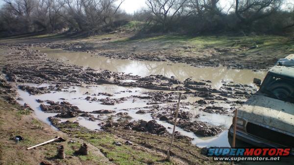 1979 Ford Bronco stuck in mud near a body of water, trees in background. Supermotors.net logo visible