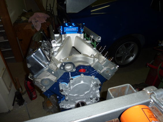 V8 engine with blue block, aluminum intake, and chrome valve covers. A blue car is visible in the background