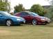 1993 Mazda MX-3 - Two Mazda MX-3s, a blue one and a maroon one, are parked side-by-side on a grassy lawn with trees in the background.