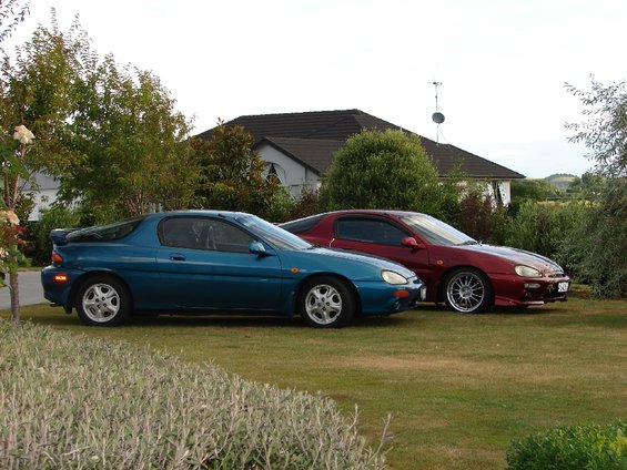 1993 Mazda MX-3 in teal and a red Mazda MX-3 parked on a lawn in front of a house
