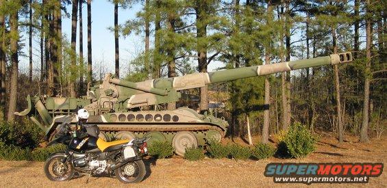 Black motorcycle with yellow seat parked next to a large artillery piece in a wooded area