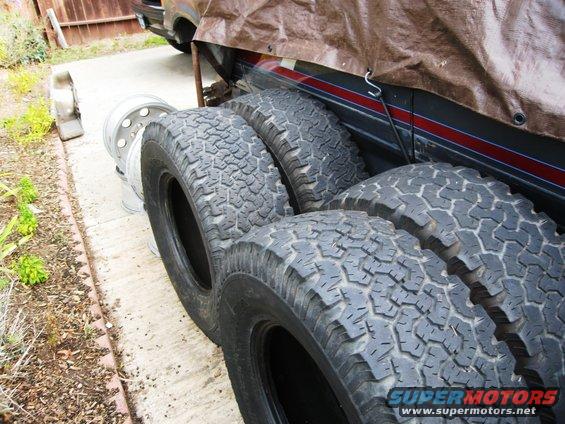 Four off-road tires and chrome wheels stacked, with a tarp-covered vehicle in the background