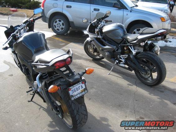 Two motorcycles parked on asphalt with a silver SUV behind them. The motorcycle on the left is black, and the one on the right is dark gray