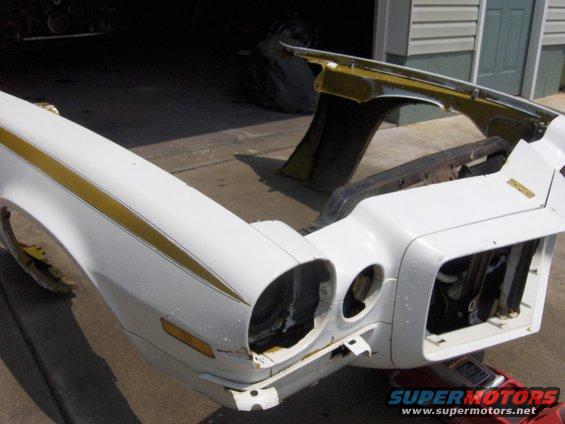 White car front end with gold stripes, missing headlights and grille. Visible rust and damage