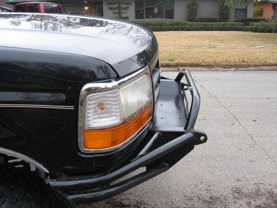 Black truck front corner with chrome trim around headlight and aftermarket steel bumper. Visible tire tread