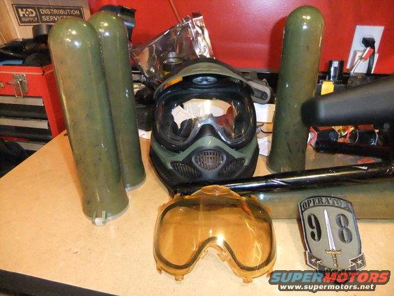 Paintball mask, spare lens, and four green air tanks on a table