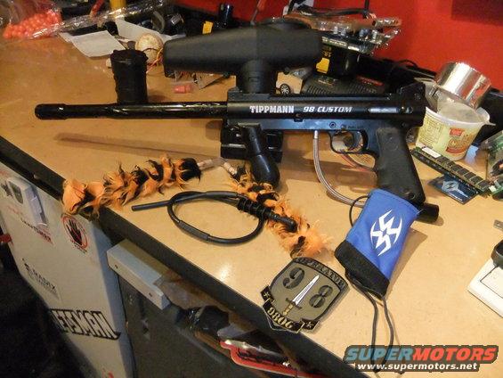 Black Tippmann 98 Custom paintball marker with orange and black feather boa and blue wristband on a wooden surface