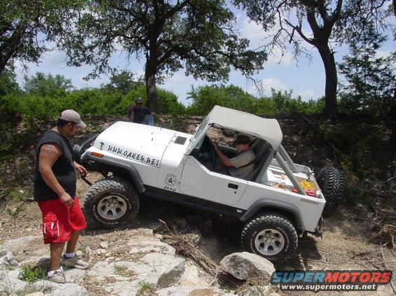 White Jeep Wrangler with a soft top on a rocky trail, driver visible. Text on hood reads WWW.CAKEN.NET. Another person stands by the front fender
