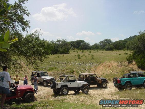 Several Jeeps parked in a grassy field with trees and hills in the background. People are standing around the vehicles