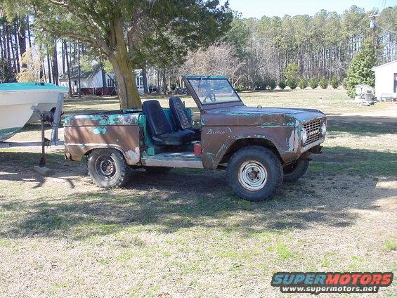 Rusty Ford Bronco with no top, sitting in a grassy yard next to a boat on a trailer