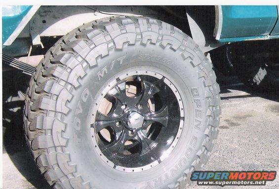 Close-up of a large, knobby off-road tire on a chrome and black aftermarket wheel, attached to a teal vehicle with visible leaf springs
