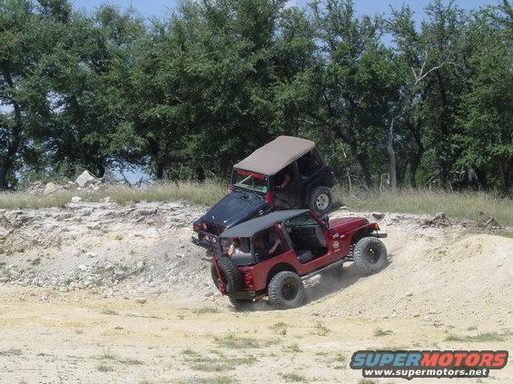 Red Jeep climbing a dirt hill with a black Jeep on top of it