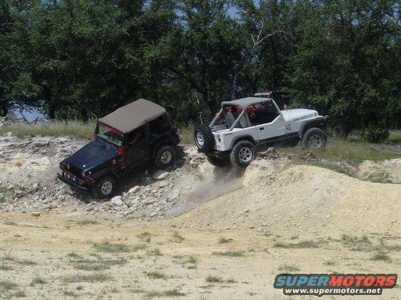 White Jeep airborne over dirt mound, black Jeep on left
