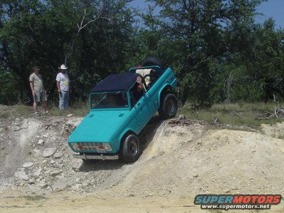 Teal Ford Bronco climbing a dirt hill, front 3/4 view. Black top, spare tire visible. Two men stand on left