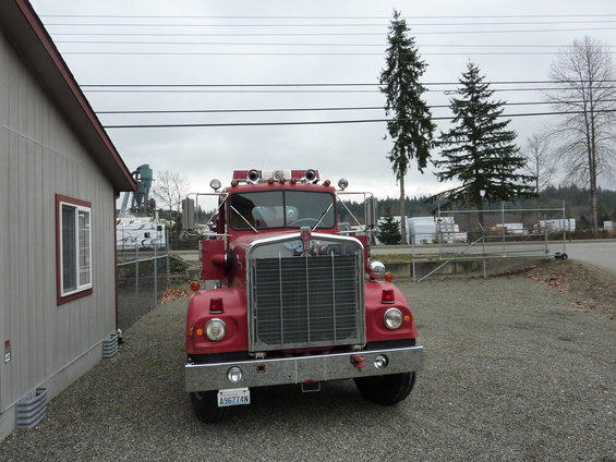 Red 1981 Kenworth W900A fire truck parked on gravel, front view. Chrome grille, round headlights, emergency lights on roof