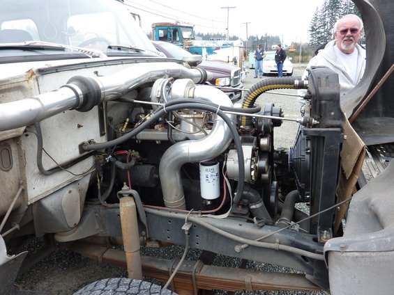 Engine bay of a truck, showing turbocharger, intake piping, oil filter, and radiator. A man stands to the right