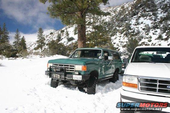 Green Ford Bronco with bull bar and winch in snow, white Ford truck next to it