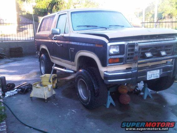 Ford Bronco on jack stands with a bucket and hose nearby. License plate reads 3JFT112