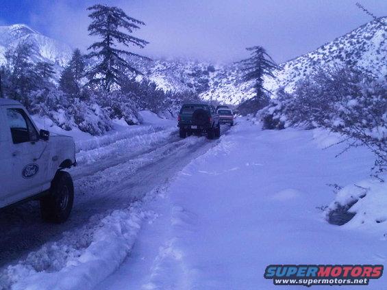 White Ford truck in foreground, two SUVs behind on snowy road, mountains and trees in background