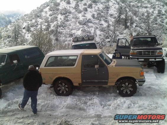 Tan and gray Ford Bronco parked in snow with other trucks nearby