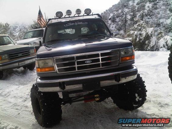 Dark blue Ford Bronco with lifted suspension and off-road tires parked in snow, another vehicle visible to the left
