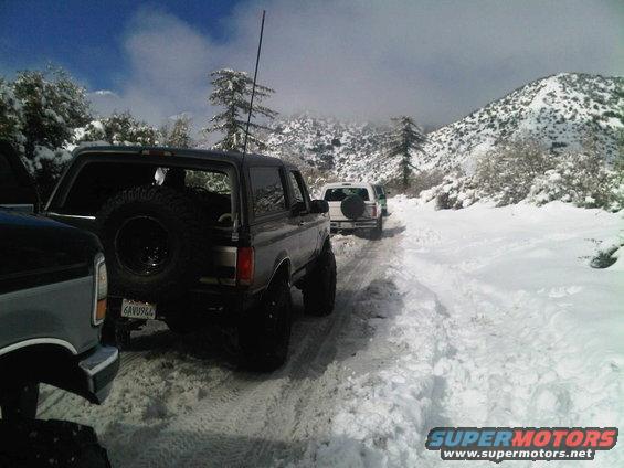 Ford Bronco with spare tire on back, driving on snowy road with other vehicles behind it