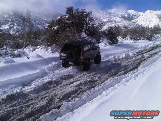 Black 1987 Ford Bronco driving on a snowy mountain road, rear view