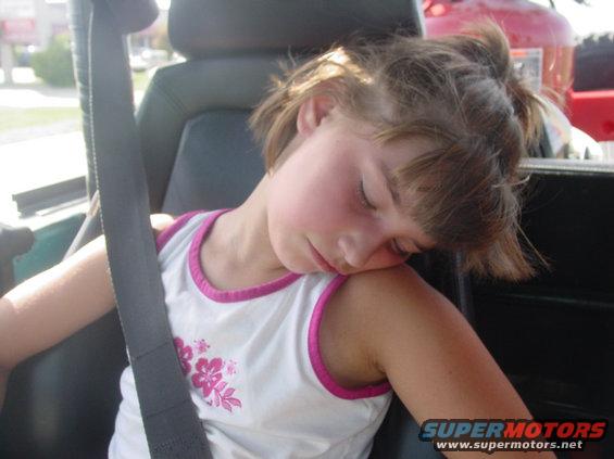 Young girl sleeping with her head on her shoulder, wearing a white tank top with pink trim and floral pattern. Seatbelt is visible
