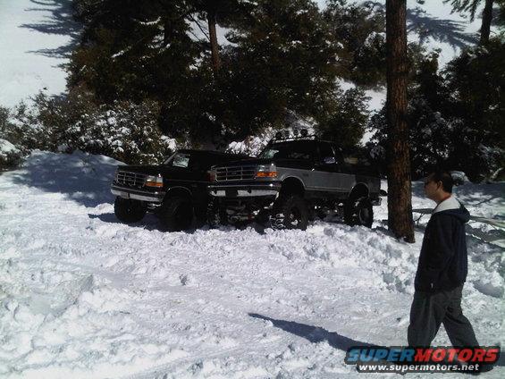 Two lifted Ford pickup trucks parked in the snow. One is black, the other is silver and black. A person walks by on the right