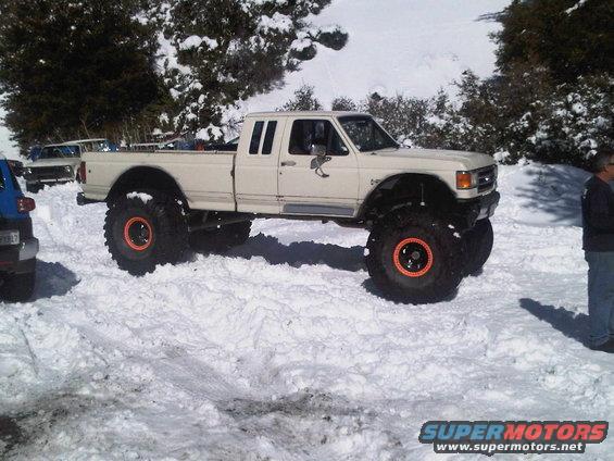 White 1987 Ford F-250 pickup truck with large off-road tires parked in snow. Another vehicle is partially visible to the left