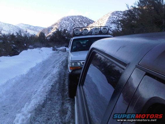 Two SUVs driving on a snow-covered mountain road. The front vehicle has a light bar on its roof. The rear vehicle is dark colored