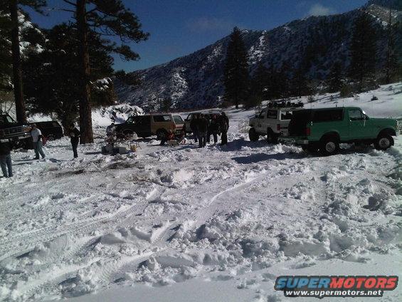 several trucks and SUVs parked in snow, some with people around them, mountains in background