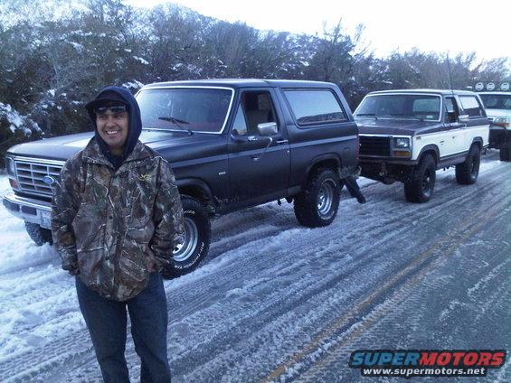Man in camo jacket stands by dark 1980s Ford Bronco; two other Broncos are behind it on a snowy road