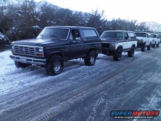 Several Ford Broncos in a line on a snowy road. The lead Bronco is dark colored, followed by a two-tone Bronco and others