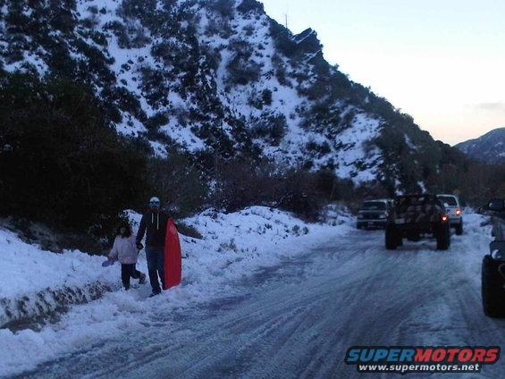 Snowy mountain road with people and sled, multiple SUVs and trucks visible