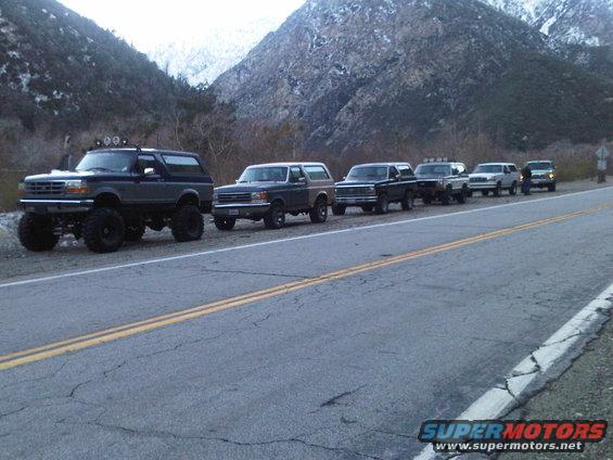 Line of lifted Ford Broncos and SUVs parked on a roadside with mountains in the background