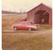1927 Dodge 1500 - A classic red and white two-door sedan with prominent fins sits in a grassy yard next to a rustic red barn.