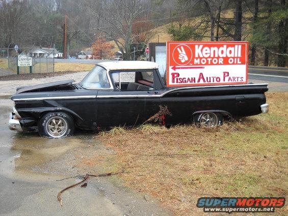 Black 1959 Ford Ranchero with a white roof, hood up, parked on grass next to a Kendall Motor Oil sign